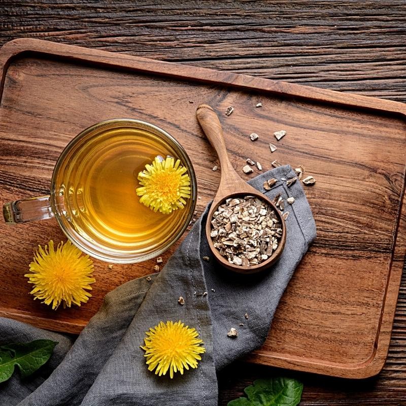 Fragmented dandelion root in a wooden spoon next to a tea and fresh flowers and leaves